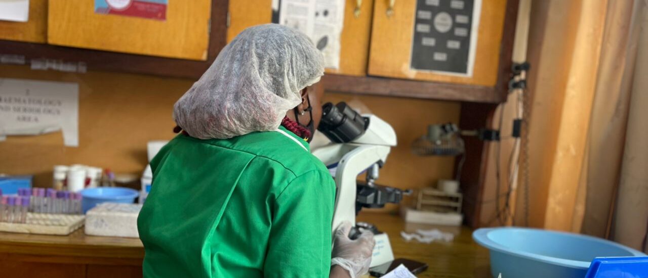 A woman siting at a microscope. She is wearing a green dress and her back is to the camera.