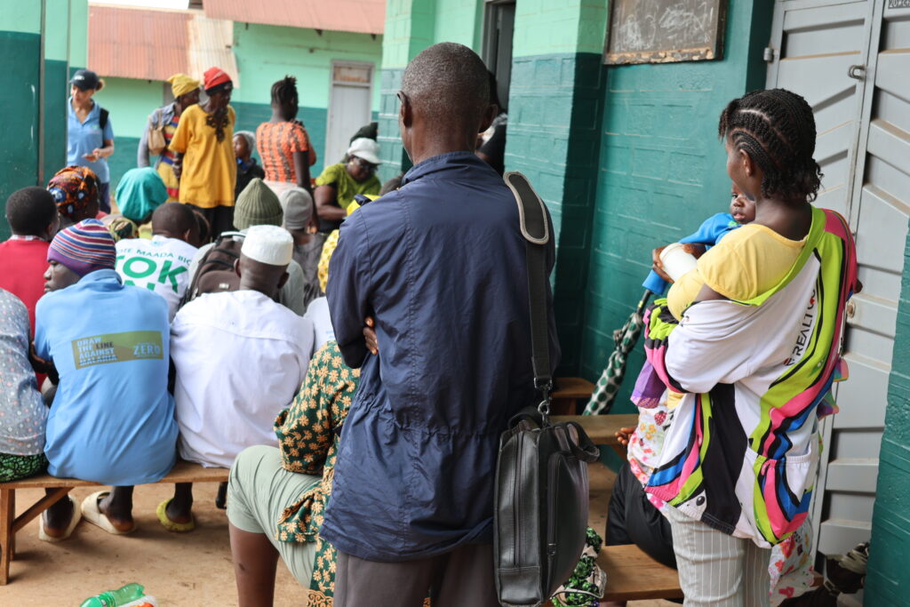 People sitting on a bench and some standing waiting to receive treatment at an epilepsy outreach